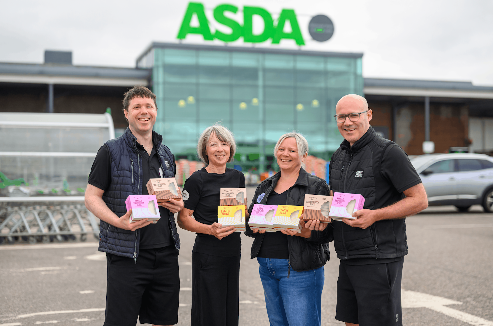The Harry Gow team standing outdoors in front of an ASDA store, each holding a sharing cake product. The background shows the ASDA building with its green logo, and a silver car is parked to the right.